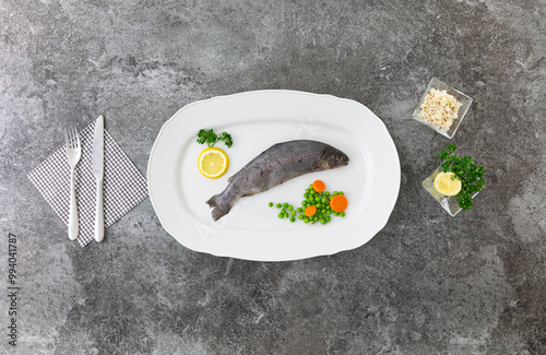 single whole rainbow trout on a white serving plate with knife and fork, lemons, parsley, peas and carrots on a grey marble table Checked chessboard napkin