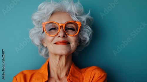 A stylish elderly woman with curly white hair, wearing oversized orange-framed glasses and an orange shirt, poses against a teal backdrop, exuding confidence. photo