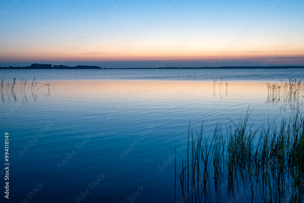 Ruhiger See bei Sonnenuntergang mit Schilf und sanften Farben im Vordergrund Ruhiger See bei Sonnenuntergang mit Schilf und sanften Farben im Vordergrund