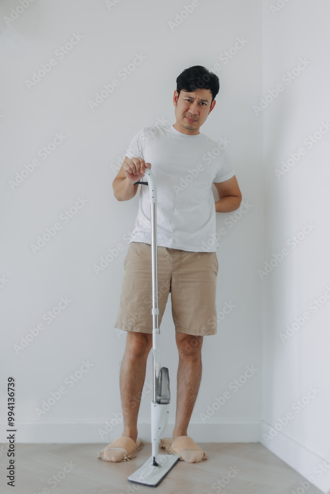 Asian man tired and painful face, standing and holding mop, hand on his waist and back, backache symptom after done housework, mopping the floor. Housework concept. Asian man tired and painful face, standing and holding mop, hand on his waist and back, backache symptom after done housework, mopping the floor. Housework concept.