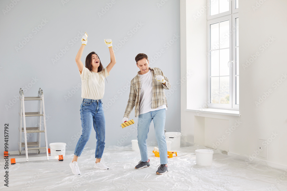 Fototapeta premium Happy young married couple have fun while doing apartment repair, painting wall with rollers. Cheerful spouses dancing together at their own new apartment, enjoying festive mood and celebrating.