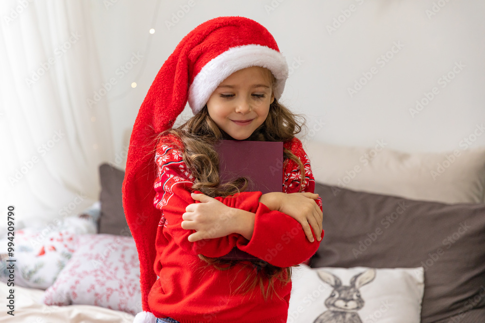 Happy little girl in red Santa hat hugging her favorite book while sitting on bed at home on Christmas morning Happy little girl in red Santa hat hugging her favorite book while sitting on bed at home on Christmas morning