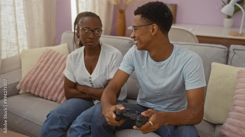 Mother crossing arms while son plays video game sitting on a couch in a cozy living room at home Mother crossing arms while son plays video game sitting on a couch in a cozy living room at home