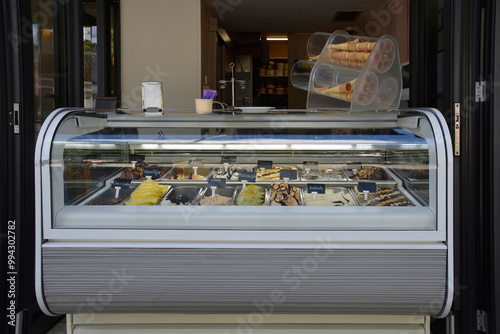 A modern ice cream parlour display case with a variety of colourful ice cream flavours. The case has a curved glass top and a sleek stainless steel base.Ice cream cones dispenser on top of the counter