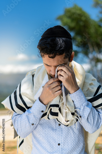 Young Jewish Man Outdoors with Tallit and Kipa, Passing Tzitzit Over His Eyes During Shacharit Prayer. vertical photo