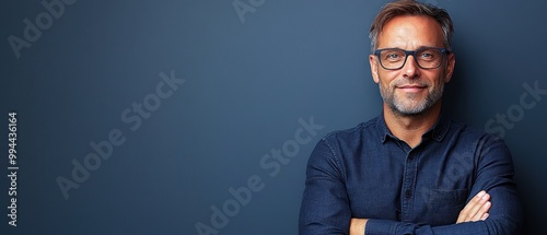 A middle-aged man wearing glasses exudes confidence with crossed arms, smiling softly against a solid blue backdrop