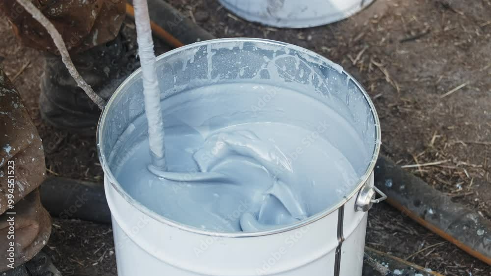 A painter working outdoors stirs silver paint in a bucket using a special tool of an electric drill before painting. Construction and repair A painter working outdoors stirs silver paint in a bucket using a special tool of an electric drill before painting. Construction and repair
