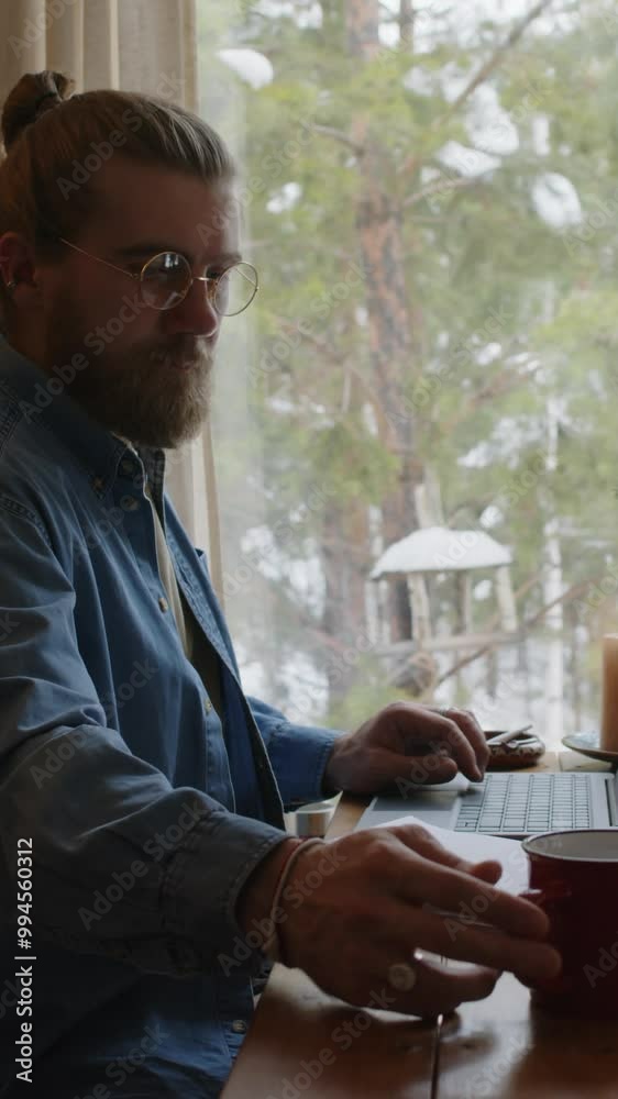 Vertical side shot of male bearded author in glasses and denim shirt working on new book, typing on laptop and drinking hot drink, looking out window at winter forest while in country house