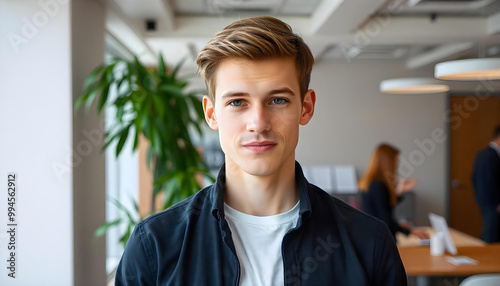 A young Caucasian man with short blond hair, wearing a navy blue jacket and a white t-shirt, looking directly at the camera with a neutral expression