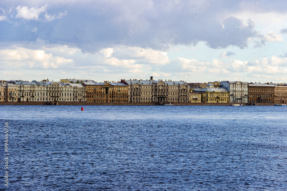 Fototapeta premium A panoramic view of the historic waterfront of St. Petersburg, Russia, showcasing rows of classic buildings along the Neva River under a partly cloudy sky.