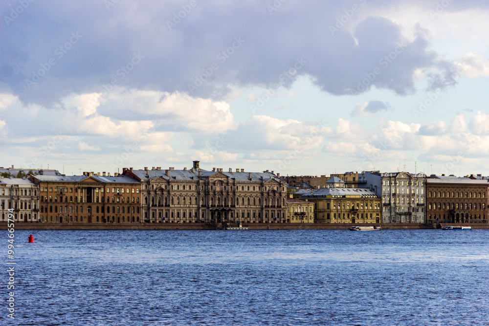 Fototapeta premium A panoramic view of the historic waterfront of St. Petersburg, Russia, showcasing rows of classic buildings along the Neva River under a partly cloudy sky.