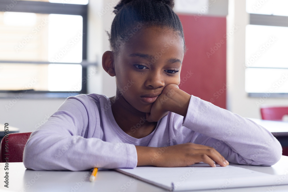 In school, african american girl sitting at desk looking at notebook, appearing thoughtful In school, african american girl sitting at desk looking at notebook, appearing thoughtful