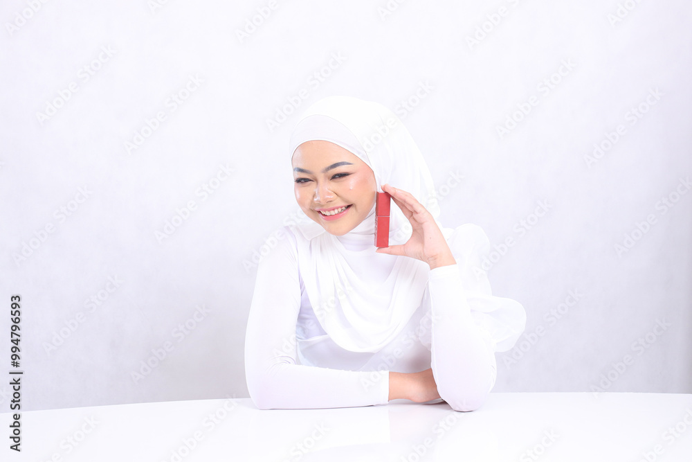 Beautiful young hijab woman indonesia happy looking at camera hand holding lipstick container and stick on table isolated white background. beauty education concept, lip care, lipstick container Beautiful young hijab woman indonesia happy looking at camera hand holding lipstick container and stick on table isolated white background. beauty education concept, lip care, lipstick container