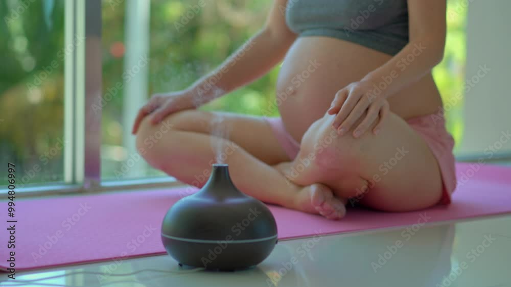Closeup of a pregnant woman meditates in front of a big sunlit window with an aroma diffuser working nearby. This serene video captures the essence of relaxation and mindfulness during pregnancy