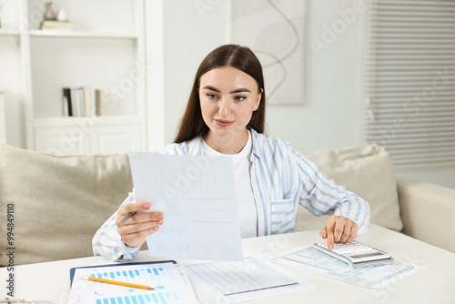 Budget planning. Beautiful young woman with papers using calculator at white table indoors photo