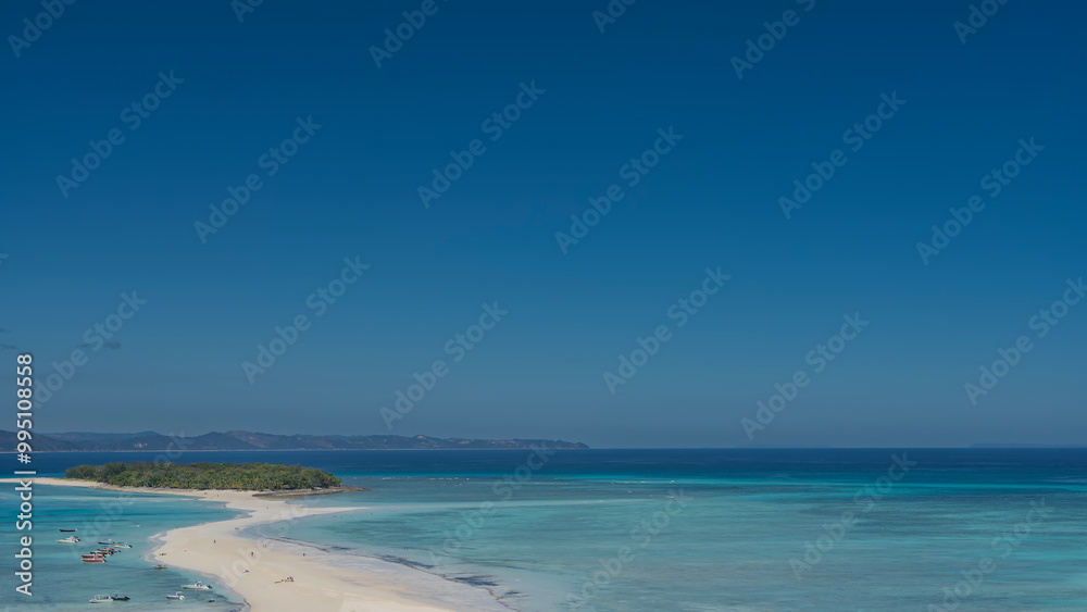 Obraz premium A delightful postcard tropical landscape. A sandy winding spit leads to a small island covered with green vegetation. Tiny silhouettes of people on the beach. Boats in the aquamarine ocean. Madagascar
