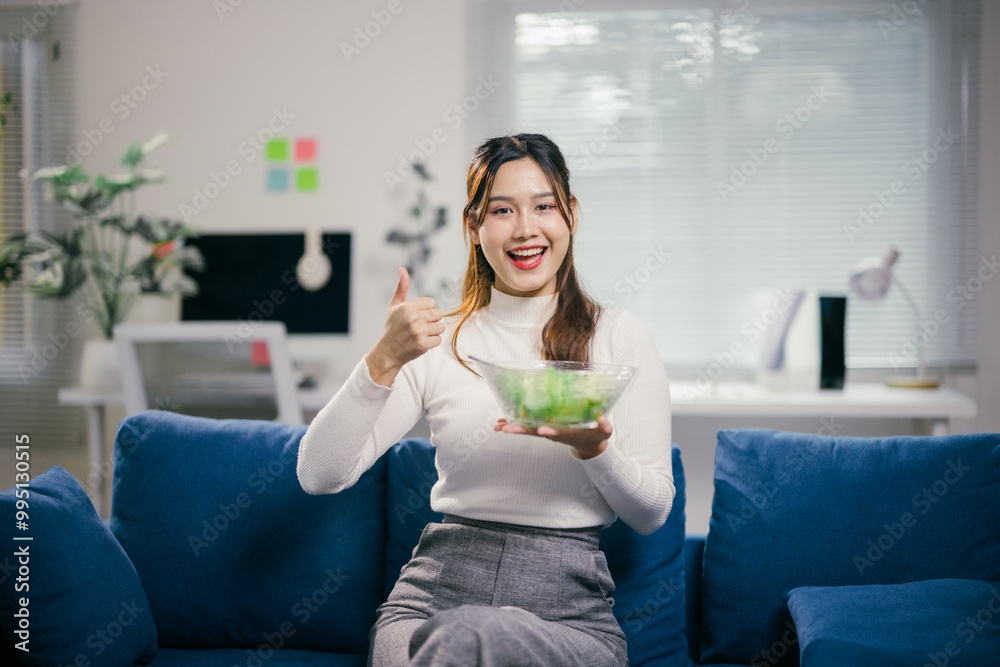 Young businesswoman is sitting on a blue couch in a modern office holding a bowl of fresh salad and showing thumbs up gesture Young businesswoman is sitting on a blue couch in a modern office holding a bowl of fresh salad and showing thumbs up gesture