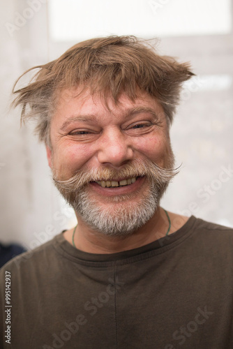 A man in a Tshirt with a beard and mustache is happily smiling for the camera, showing off his facial hair. His jawline and throat are visible, with a few wrinkles around his eyes