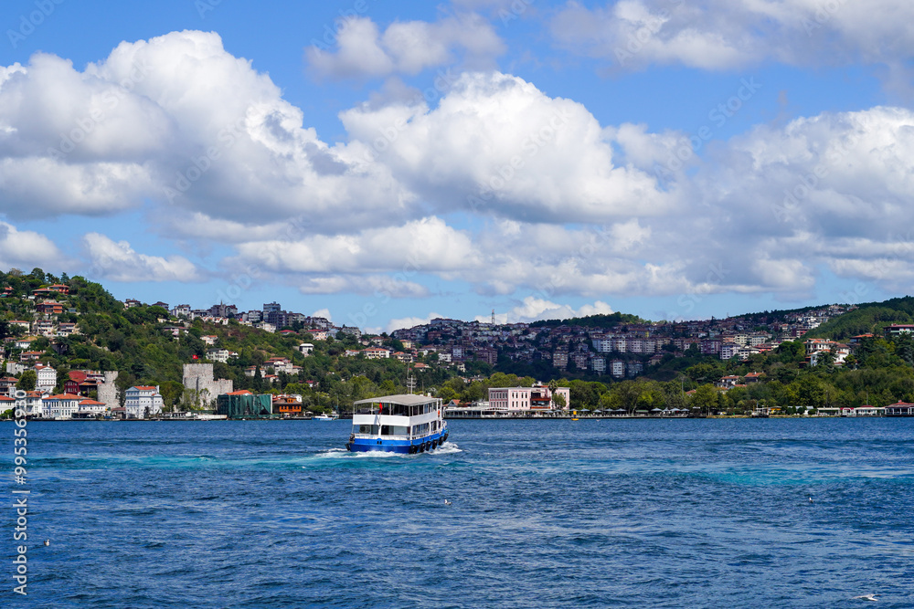 Obraz premium Passenger ferry navigating the Bosphorus with scenic views of Istanbul\'s hills and coastline under a bright, partly cloudy sky