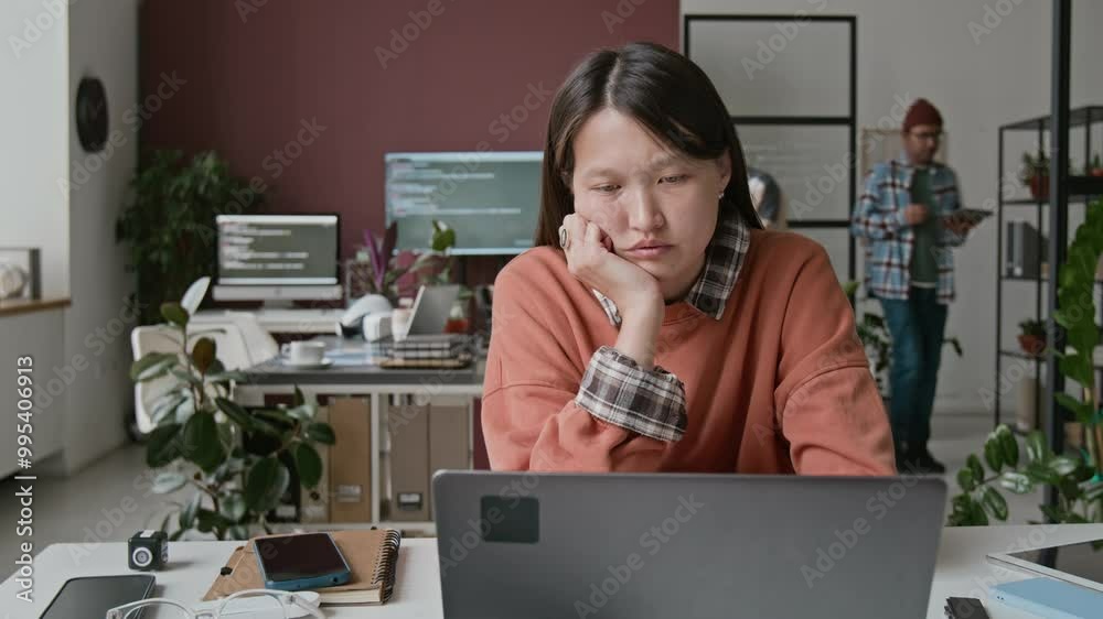 Medium timelapse shot of sleepy, burnt-out young Asian female IT engineer sitting at desk at work in busy office, staring at laptop screen with code with apathy, unable to fix bug in code