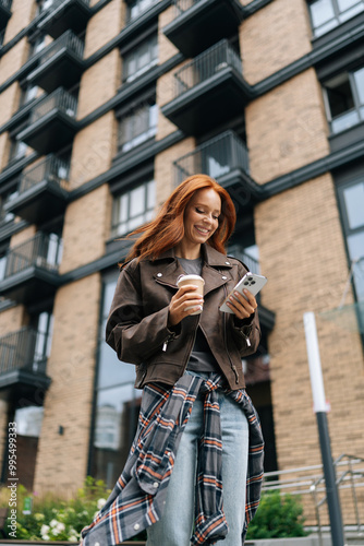 Vertical low-angle view of charming woman standing in city street, holding coffee and scrolling on phone, blending tech and fashion effortlessly. Cheerful redhead female typing smartphone outdoors