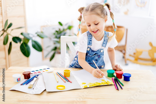 a little child girl paints a drawing with a brush in an album and smiles, a happy child draws a gouache picture at home at the table photo