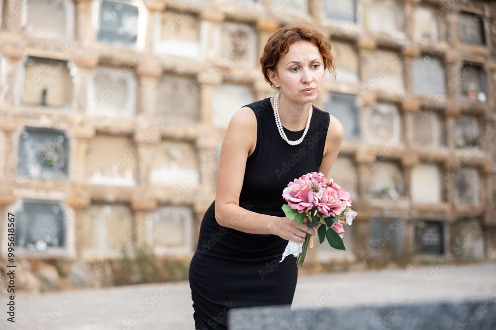 Fototapeta premium Woman in mourning wearing black dress gently resting pink bouquet on grave with sorrowful expression showing quiet reflection and deep loss in cemetery setting