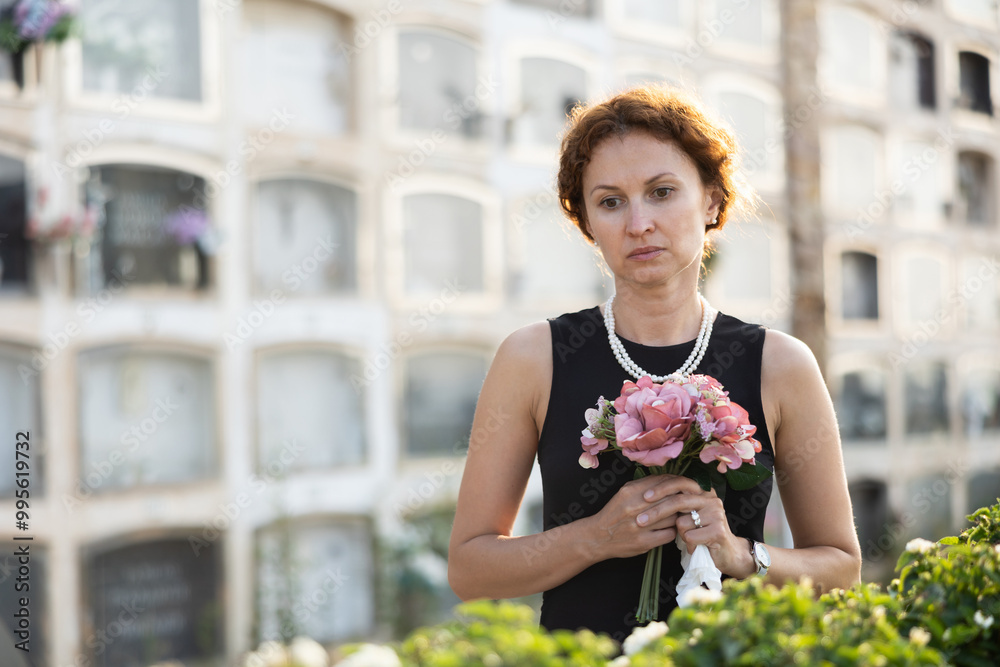 Fototapeta premium Sorrowful middle aged woman in black dress standing in cemetery holding bouquet of flowers with reflective and somber expression against backdrop of stone memorial walls
