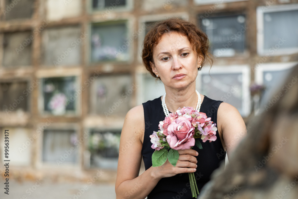 Fototapeta premium Middle aged woman wearing black dress and pearls holding bouquet of pink flowers, gazing sorrowfully at grave, reflecting deeply on loss in quiet cemetery