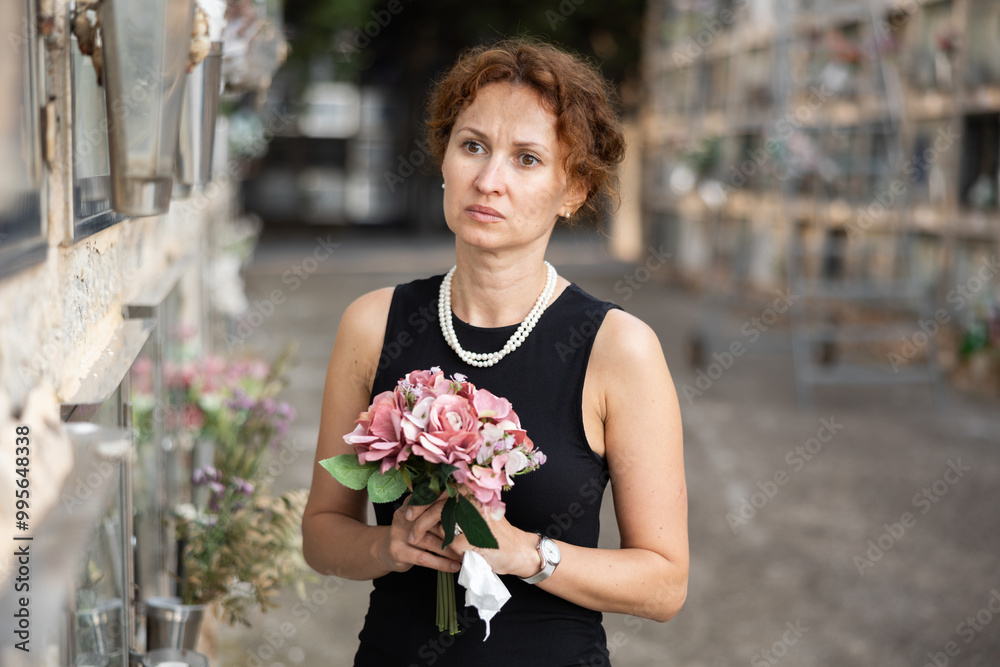 Fototapeta premium Elegant grieving woman in forties, wearing formal black dress and pearls, standing with bouquet in hands near stone columbarium wall in cemetery, gazing sorrowfully and contemplating loss ..