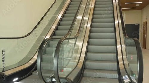 Footage shows an empty moving escalator in a supermarket or a mall shop with no people there. The escalator has rubber and glass railings and a store on the second floor. photo