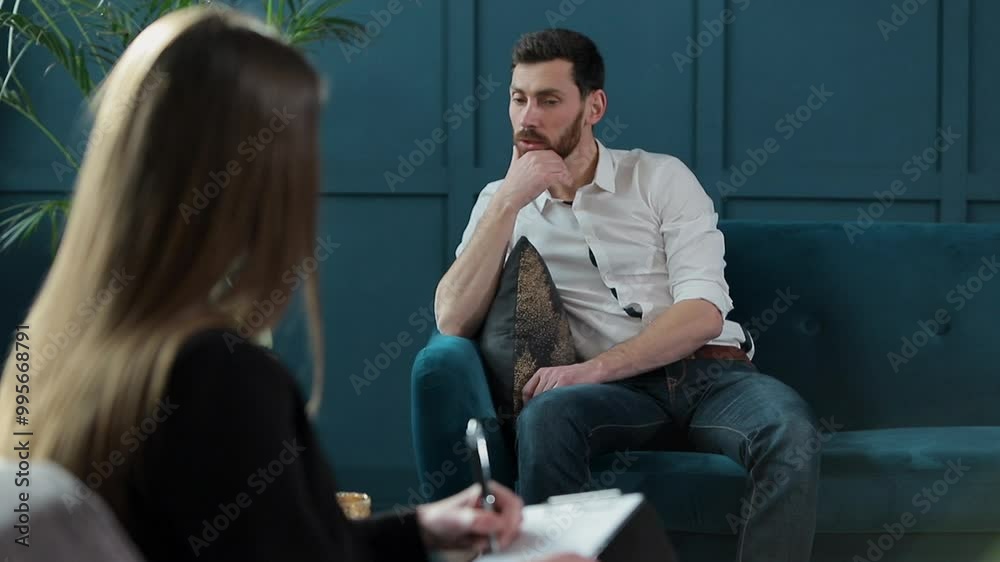 Man client sitting with female psychologist on the comfortable couch during the psychological session at the blue office interior.