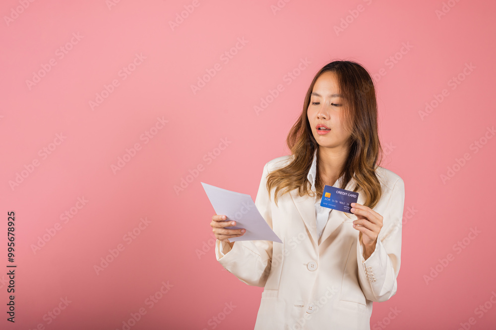 Portrait of Asian young woman holding paper calculating bills on hand and credit card for payment, female person problem she hold worried over bill, studio shot isolated on pink background Portrait of Asian young woman holding paper calculating bills on hand and credit card for payment, female person problem she hold worried over bill, studio shot isolated on pink background