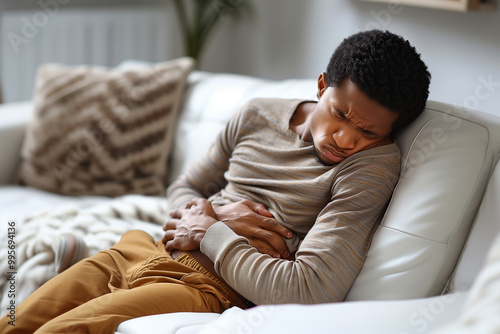 Sad young man suffering from PMS and menstruation pain at home, upset eatsern male having stomach ache, acute abdominal pain and headache, sitting on couch in living room, copy space photo