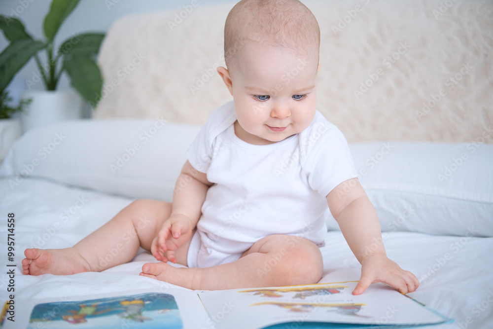 Happy funny bald baby in white bodysuit reading book sitting on parents bed Happy funny bald baby in white bodysuit reading book sitting on parents bed
