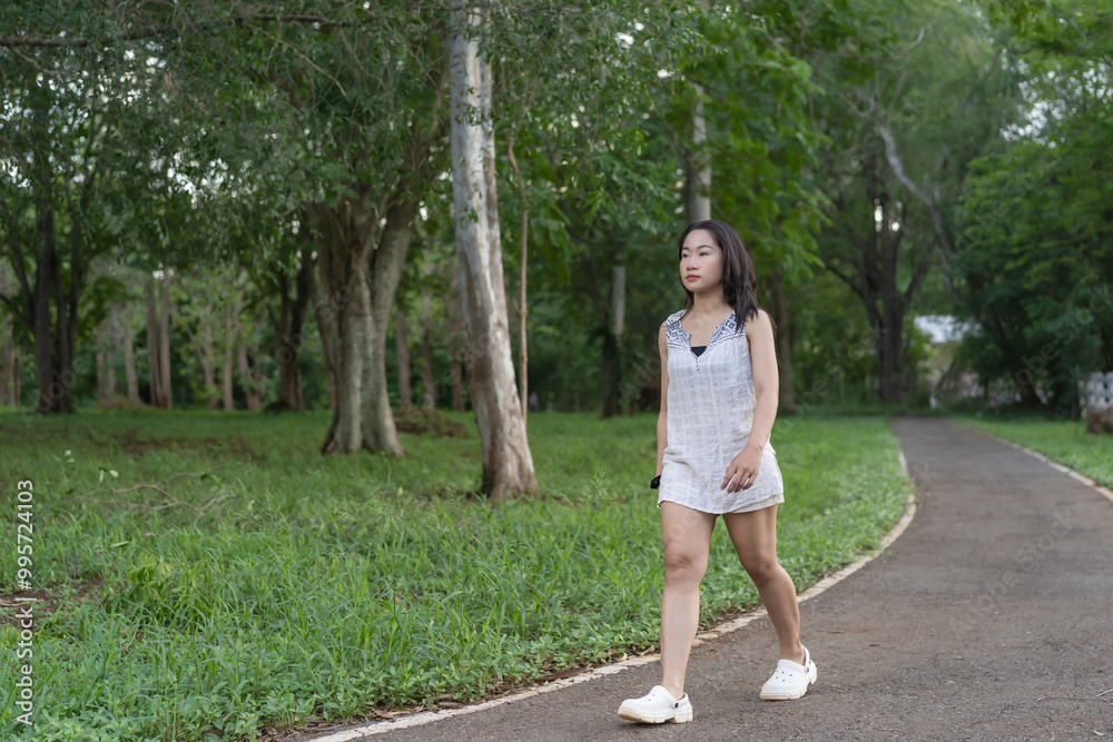 Enjoying nature young woman walking on the sidewalk in the park. Exercise by walking, a healthy, positive mood, happy steps, feeling relaxed without worry, and starting a new day. Concept of vitality. Enjoying nature young woman walking on the sidewalk in the park. Exercise by walking, a healthy, positive mood, happy steps, feeling relaxed without worry, and starting a new day. Concept of vitality.