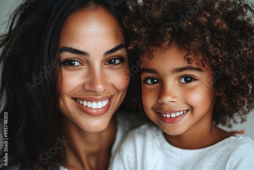 Mixed racial young mother and cute daughter smiling and interacting in front of a plain background, dressed in simple clothes, captured in a family photo portrait shoot showcasing their joyful bond