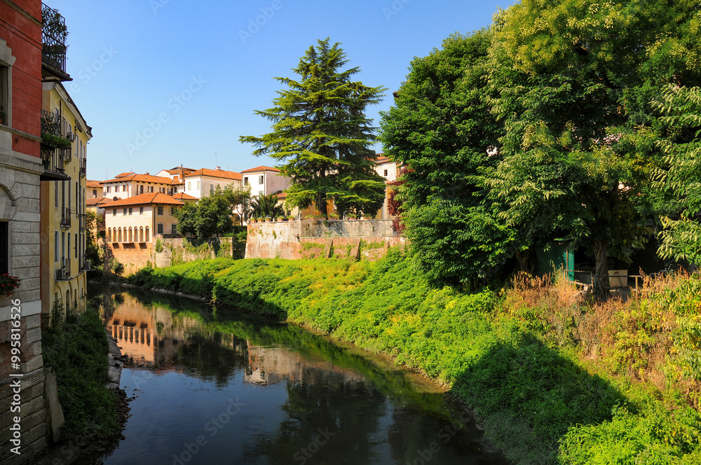Picturesque view of colorful Italian buildings along a serene river in Vicenza. Lush greenery and trees line the banks, with reflections of architecture in the calm water Picturesque view of colorful Italian buildings along a serene river in Vicenza. Lush greenery and trees line the banks, with reflections of architecture in the calm water