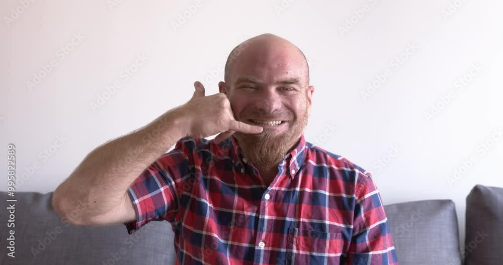 bald man with a red goatee sitting on the couch at home. He makes a call me gesture with his hand, suggesting he wants to be contacted. bald man with a red goatee sitting on the couch at home. He makes a call me gesture with his hand, suggesting he wants to be contacted.
