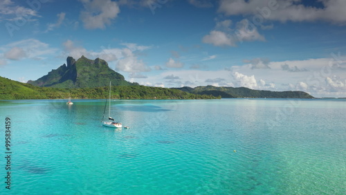 Two sailboats yachts anchored in turquoise waters coast of Bora Bora lagoon, green mountains rising under bright blue sky. Remote wild nature paradise, exotic summer luxury travel. Drone aerial flight