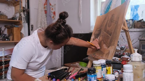 A young woman with Down syndrome is absorbed in drawing on a canvas easel in an art studio.