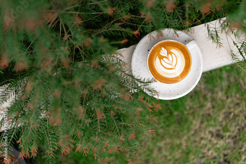 A warm cup of latte art rests on a table surrounded by green foliage in a tranquil outdoor café setting
