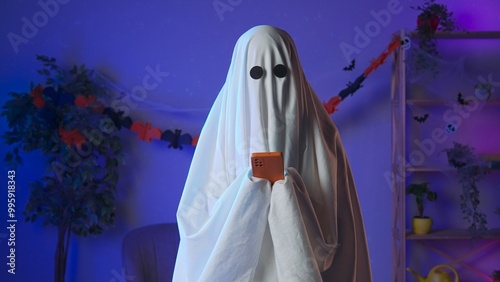 Photos Funny halloween ghost in white sheet with black eyes in dark room texting on smartphone, sending message in app