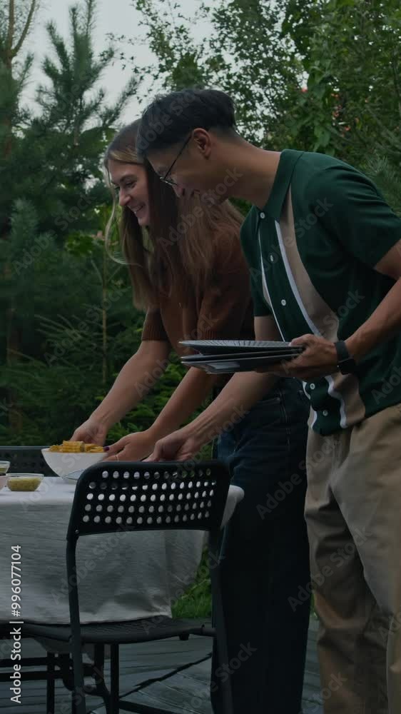 Vertical medium shot of young ethnically diverse friends chatting and setting small cozy dinner table on patio backyard while getting together in countryside on summer evening