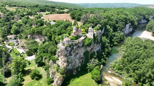 A breathtaking aerial view of a historic village nestled in lush green hills. The scene features a beautiful castle  on a cliffside, overlooking a river flowing through the valley. 
