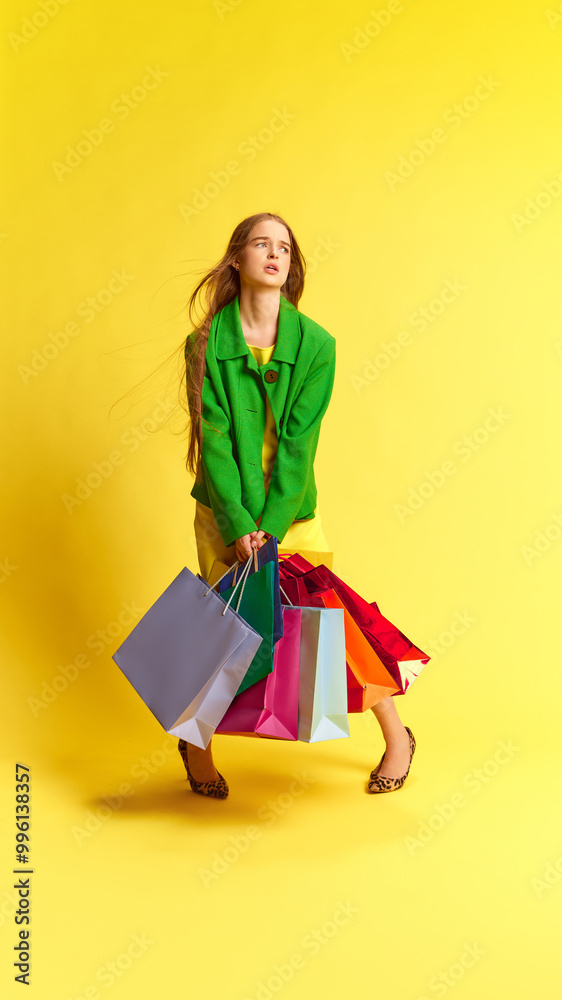Young stylish woman feeling tired while carrying many multicolored shopping bags against yellow studio background. Concept of shopping, sales, Black Friday, Cyber Monday Young stylish woman feeling tired while carrying many multicolored shopping bags against yellow studio background. Concept of shopping, sales, Black Friday, Cyber Monday