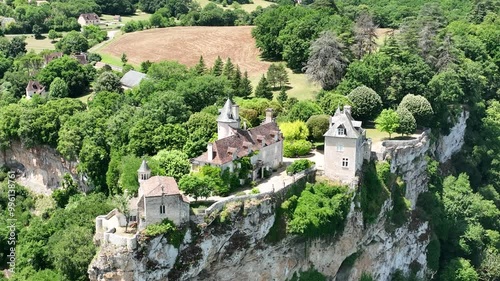 A breathtaking aerial view of a historic village nestled in lush green hills. The scene features a beautiful castle  on a cliffside, overlooking a river flowing through the valley. 