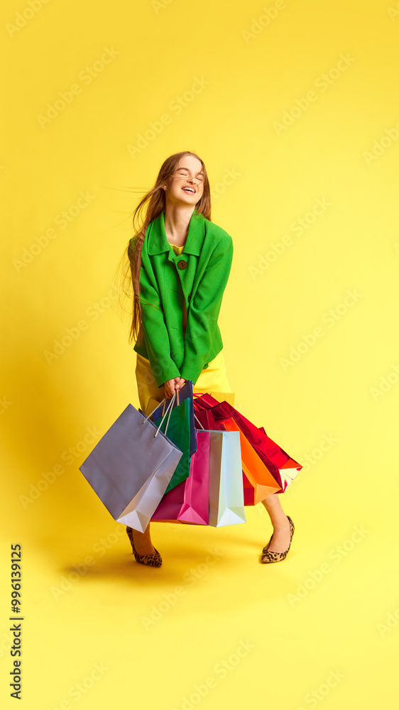 Full-length portrait of happy young woman smiling, standing with many multicolored shopping bags against yellow studio background. Concept of shopping, sales, Black Friday, Cyber Monday Full-length portrait of happy young woman smiling, standing with many multicolored shopping bags against yellow studio background. Concept of shopping, sales, Black Friday, Cyber Monday