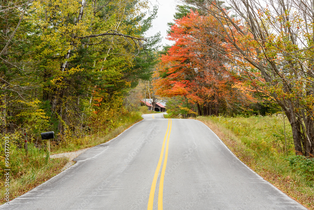 Deserted uneven curvy road lined with colourful trees in the countryside of Vermont in autumn. A wooden barn by the road is visible in distance. Deserted uneven curvy road lined with colourful trees in the countryside of Vermont in autumn. A wooden barn by the road is visible in distance.