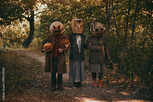 Фотография Three strange people in terrifying masks and odd clothes standing in autumn woods, creating creepy Halloween mystery